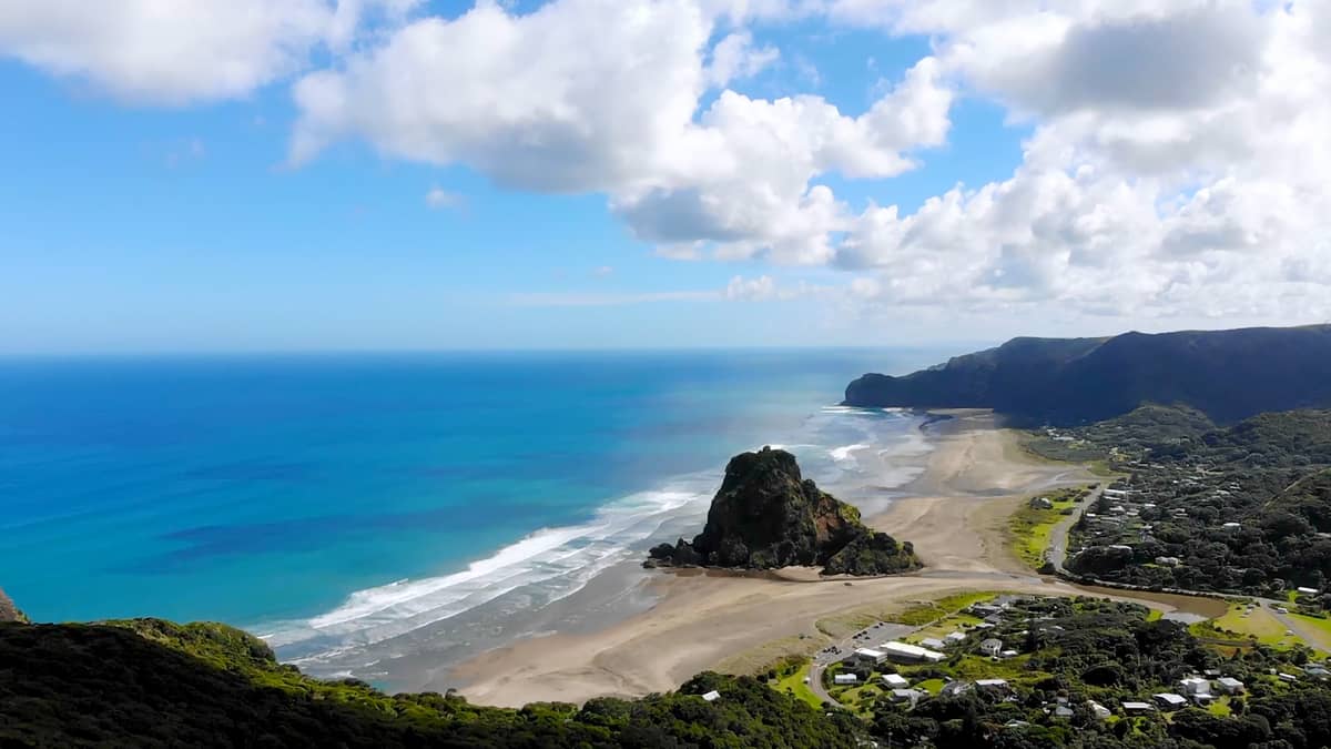 Vue aérienne de Piha Beach sur la côte ouest d'Auckland montrant une spectaculaire plage de sable noir, l'emblématique Lion Rock, des vagues océaniques et des collines verdoyantes.