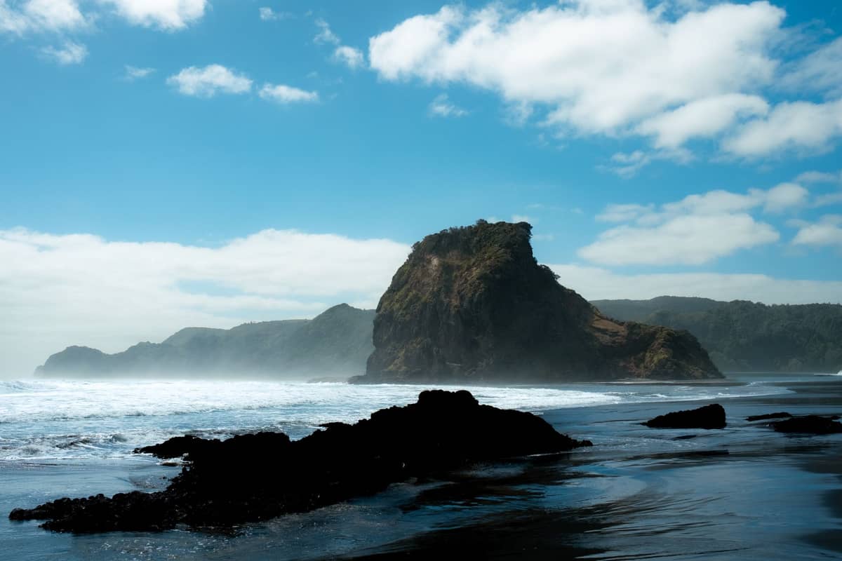 Dramatischer Schwarzsandstrand in Piha mit dem Lion Rock, brechenden Wellen und blauem Himmel mit vereinzelten Wolken an Aucklands Westküste.