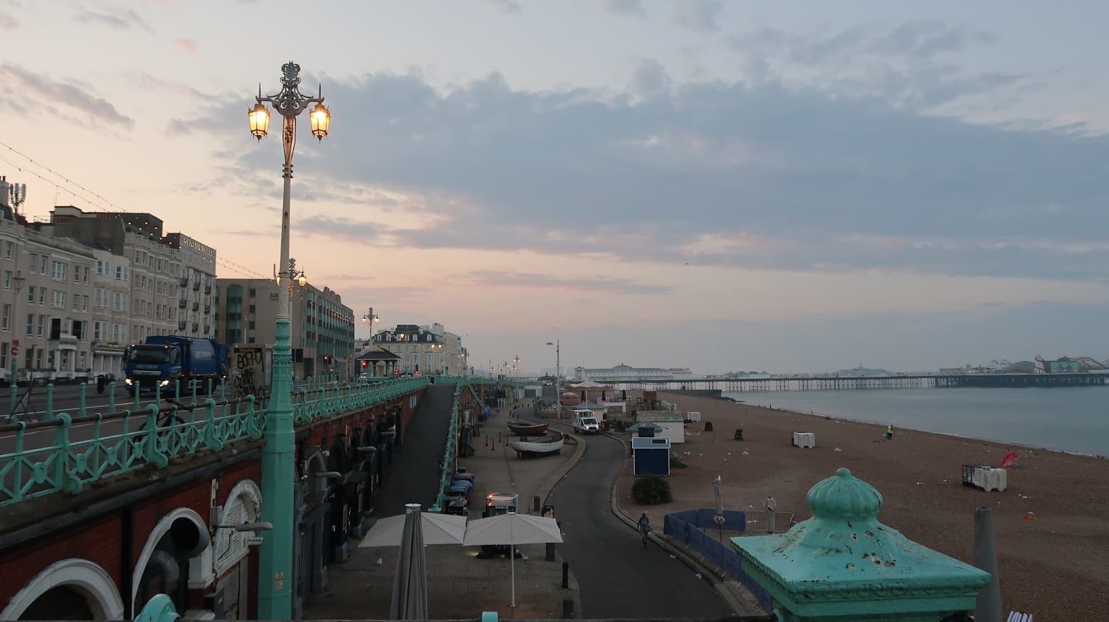 Brighton sahil cephesi, gün batımında promenad, sahil binaları, fenerler, plaj ve iskelelerle yumuşak aydınlatma ve geniş kıyı manzarasıyla gösteriliyor.