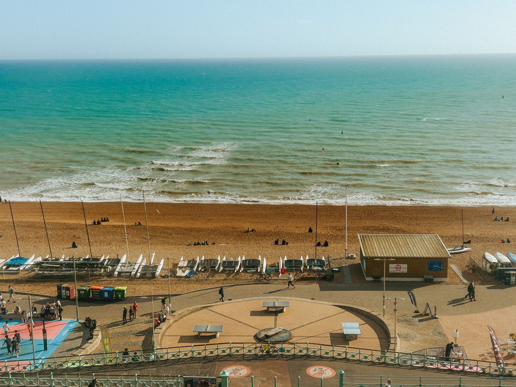Parlak bir günde Brighton'ın ana plajı ve sahil şeridinin çakıl kıyısı, plaj kulübeleri, insanlar ve denizi gösteren görünümü.