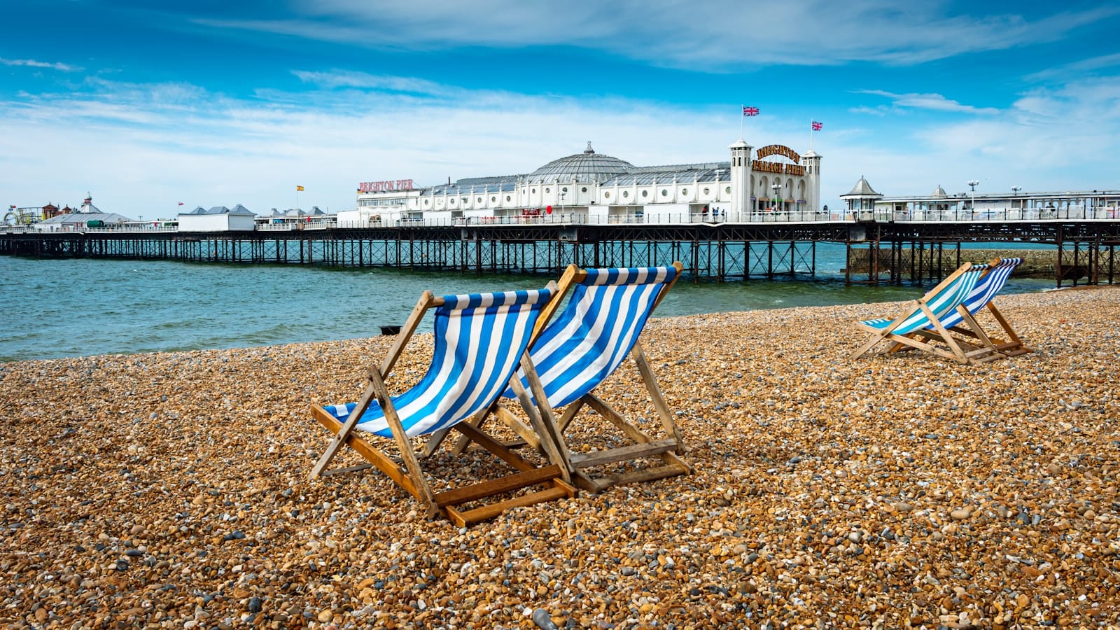 Striped deck chairs on Brighton's pebble beach with Brighton Pier and blue sky in the background, capturing a classic seaside atmosphere.