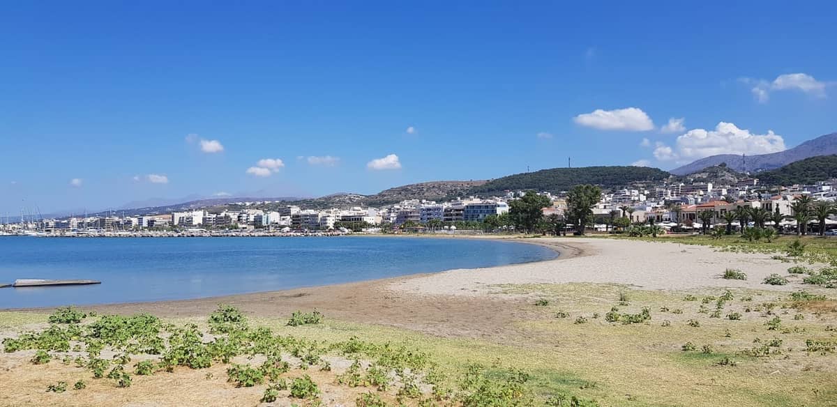 Rethymno, Girit'te açık mavi bir gökyüzünün altında berrak sulu geniş kumsal, şehir binaları ve arka planda tepeler.