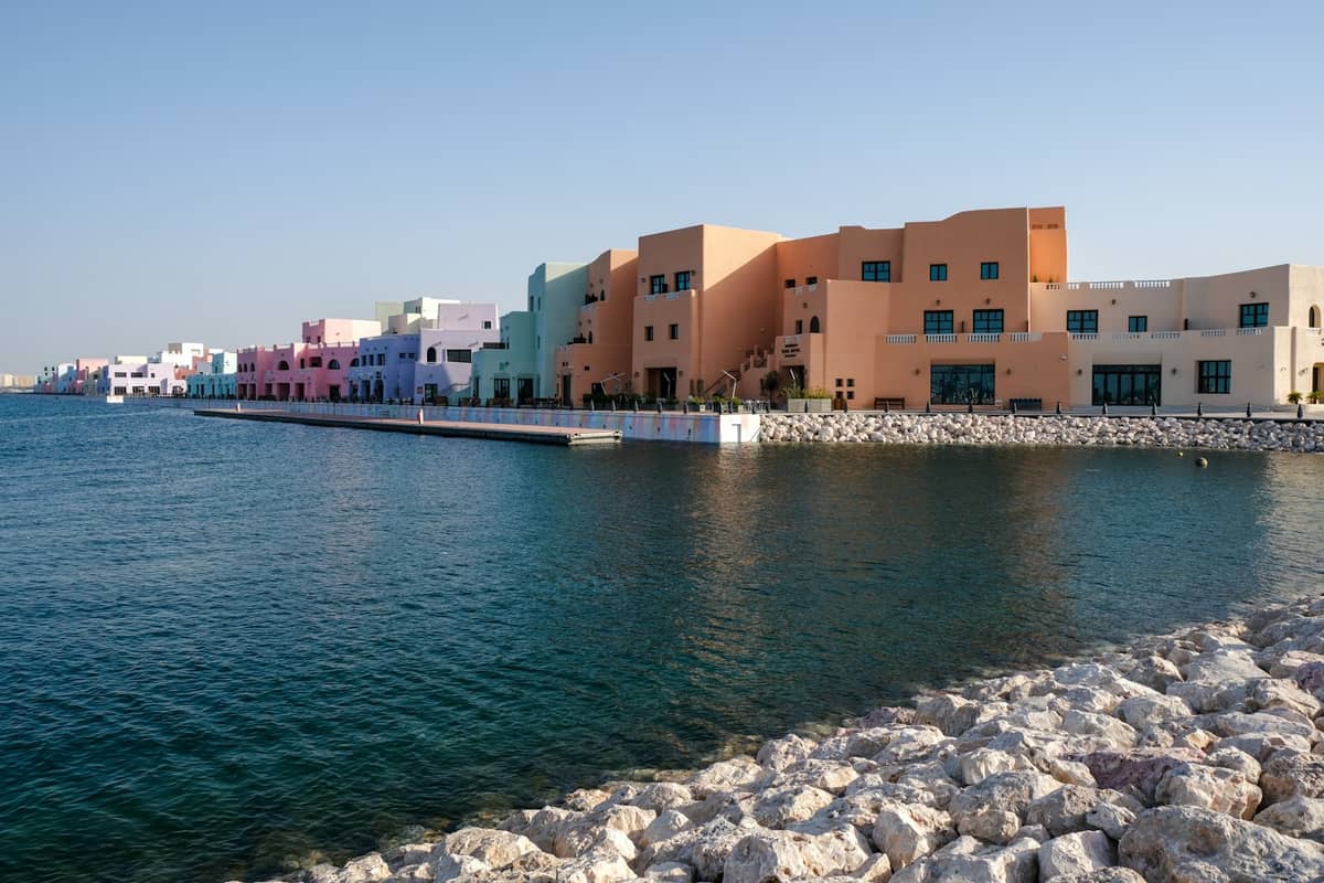 Pastellfarbene Gebäude des Katara Beach in Doha säumen die Strandpromenade, mit klarem blauen Wasser im Vordergrund und einem felsigen Ufer.