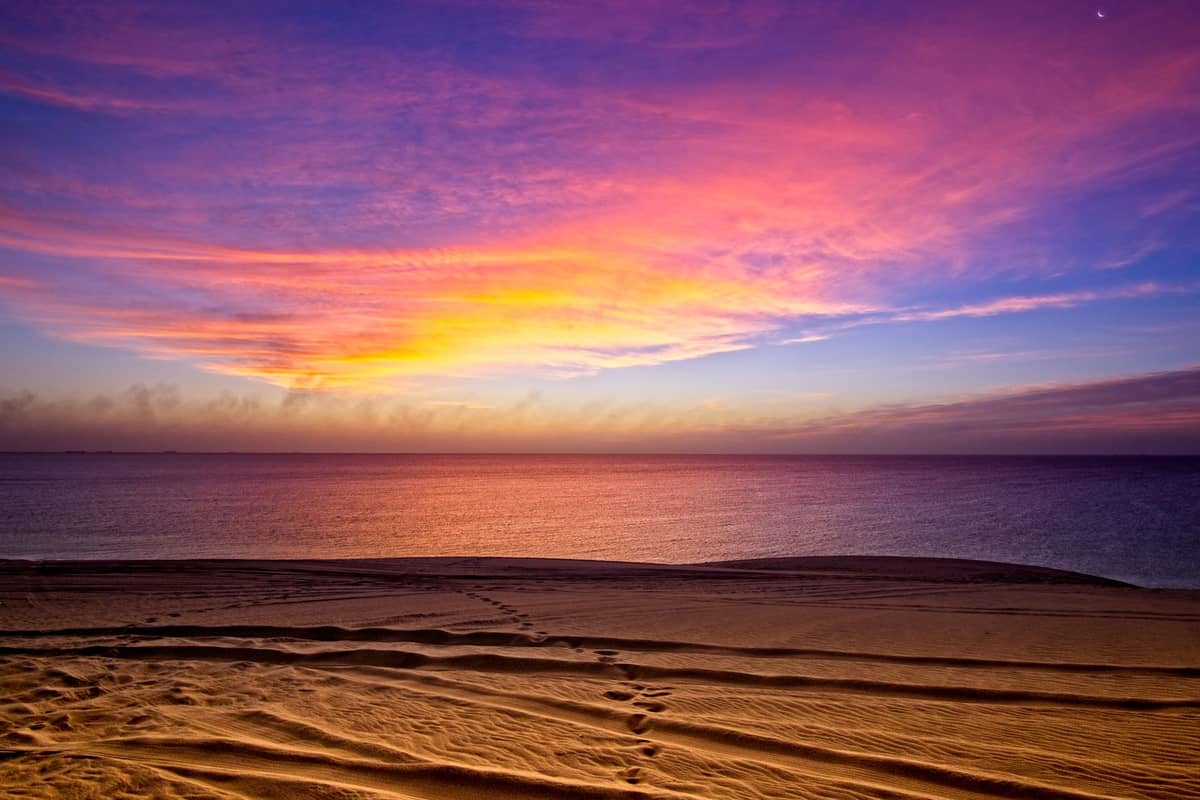 Ein dramatischer Sonnenuntergang über dem Meer mit goldenen Sanddünen im Vordergrund, die auf das ruhige Wasser unter einem farbenfrohen Himmel treffen.