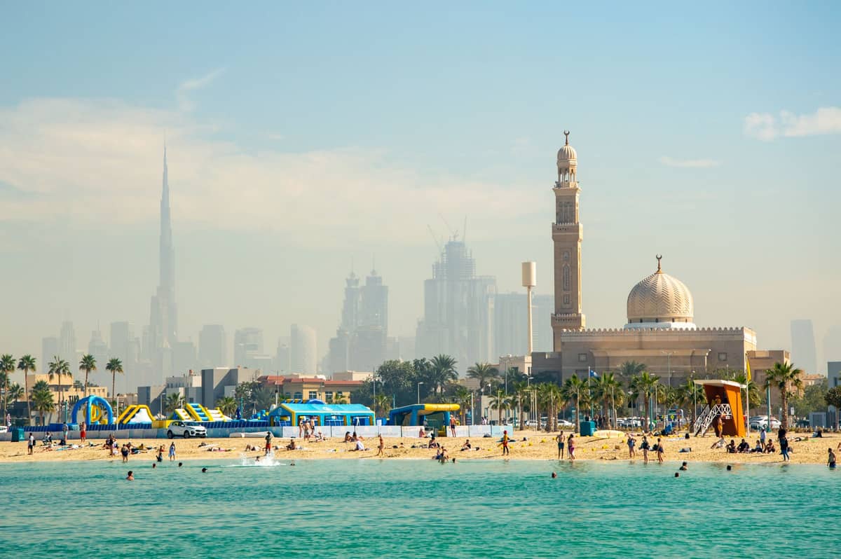 Belebter Sandstrand in Dubai mit Moschee, Palmen und dem Burj Khalifa im Hintergrund, mit Strandparkeinrichtungen und Stadtsilhouette.