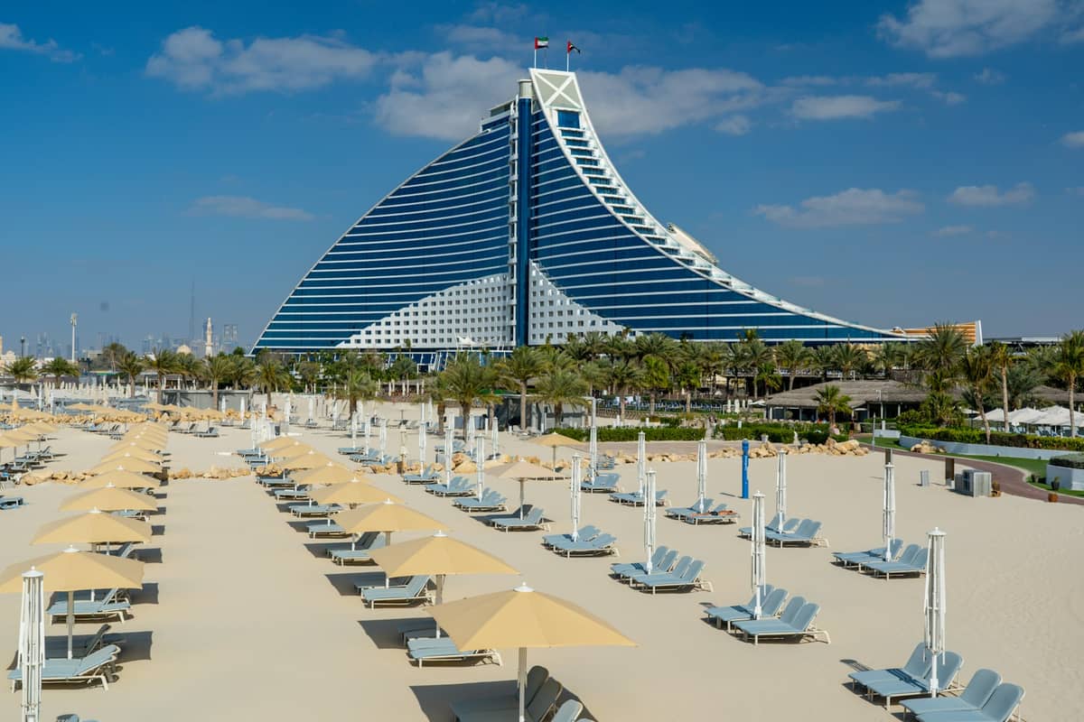 Reihen von Sonnenliegen und Sonnenschirmen an einem Sandstrand mit dem Jumeirah Beach Hotel in Dubai im Hintergrund.