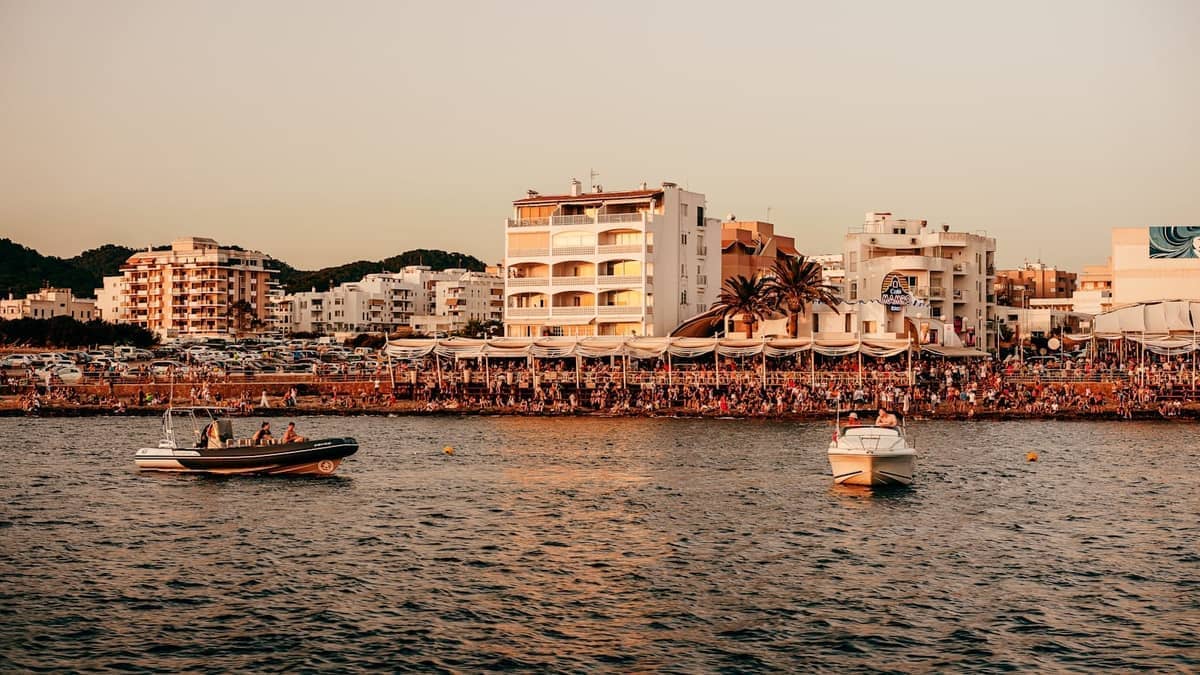 Beachfront scene in Ibiza with lively crowds, numerous bars and restaurants, and boats anchored just offshore at sunset.