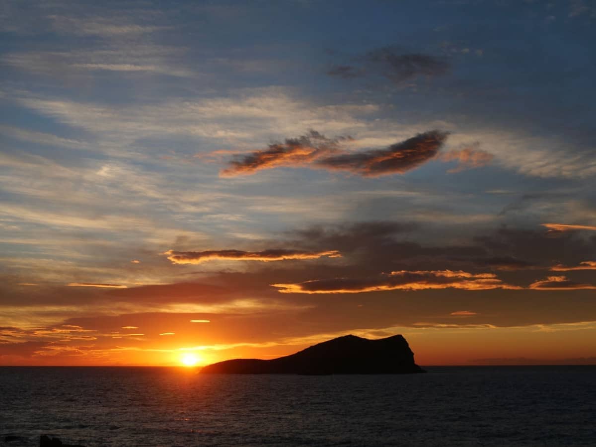 Dramatic sunset with vibrant clouds over the sea, silhouette of a rocky island on Ibiza's west coast, golden light reflecting on the water.