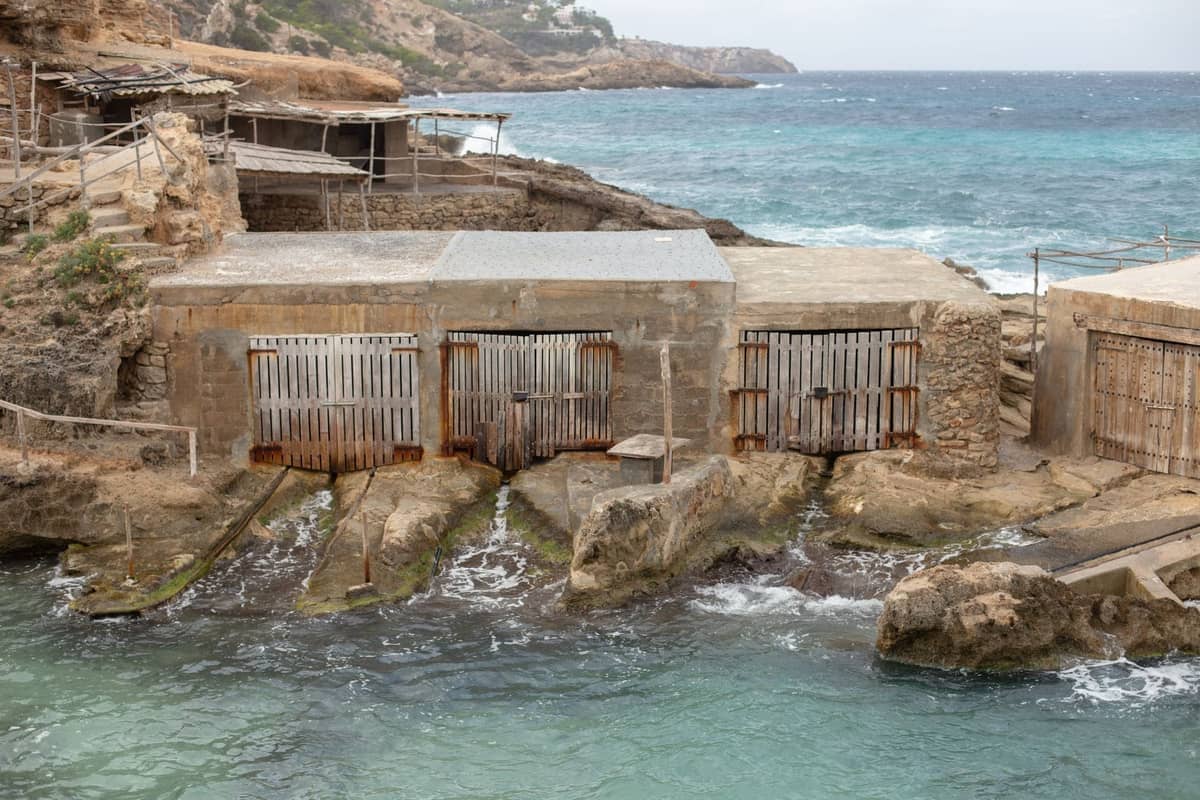 Traditional stone fisherman’s huts on a rocky coastline next to turquoise waters under a bright sky, evoking Ibiza’s unpolished northern beach character.