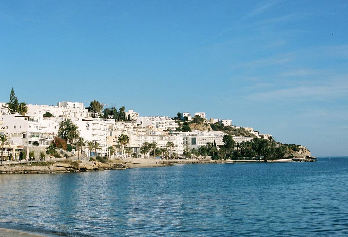 View of whitewashed buildings along the coastline in Ibiza Town with calm blue sea under clear sky.
