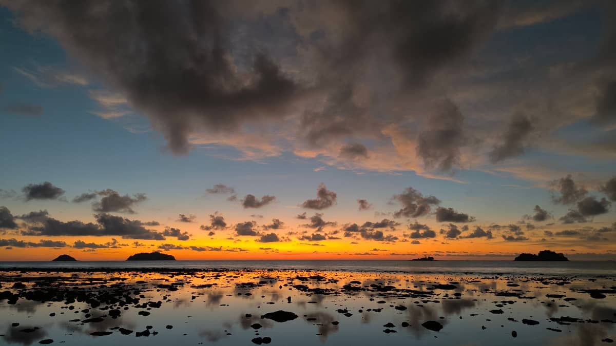 Ein ruhiger Strand bei Sonnenuntergang auf Koh Lanta, mit verstreuten Felsen auf nassem Sand und mehreren kleinen Inseln als Silhouette vor dem farbenprächtigen Himmel.