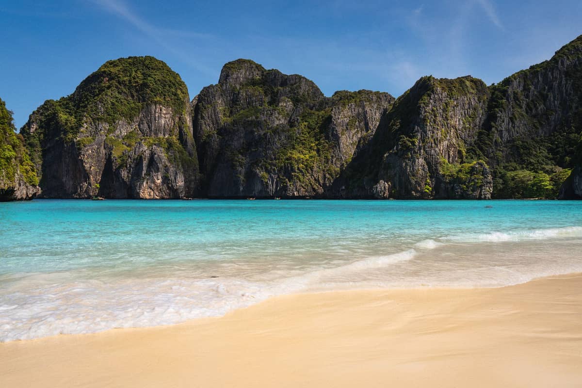 Sandstrand mit türkisfarbenem Wasser und dramatischen Kalksteinfelsen, typisch für die nur per Tour erreichbaren Inselstrände von Krabi unter blauem Himmel.