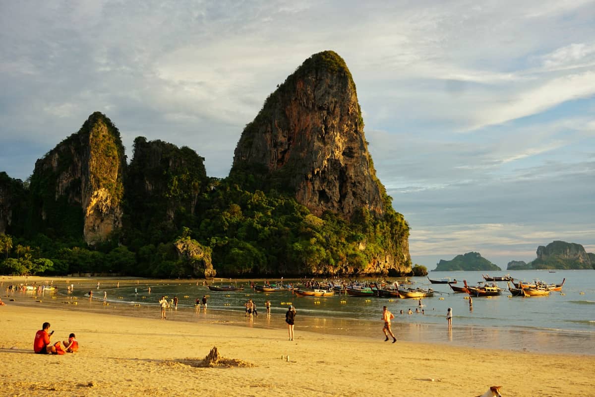 Breiter Sandstrand mit Menschen, Longtail-Booten und beeindruckenden Kalksteinfelsen auf der Halbinsel Railay in Krabi, Thailand.