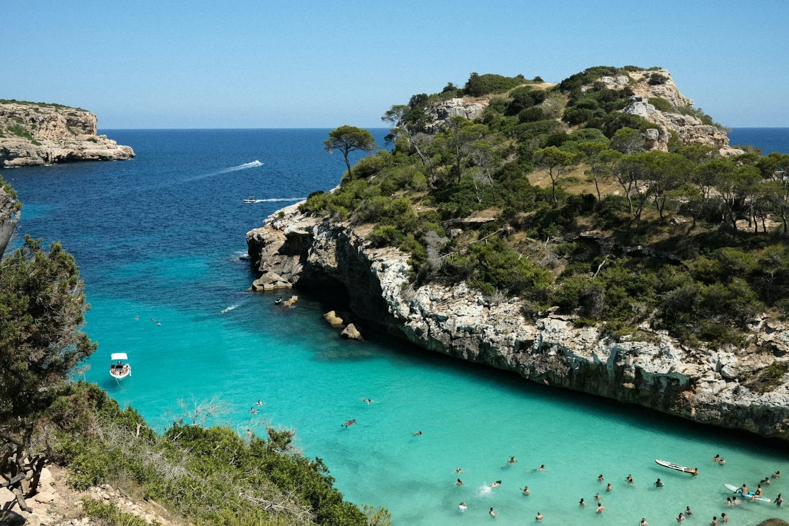 Enseada rochosa e dramática em Maiorca com água turquesa, banhistas, barcos e colinas verdes sob um céu azul límpido. Cena de praia perfeita e convidativa.