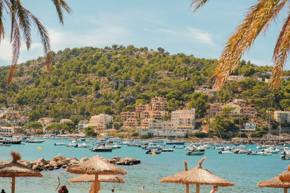Blick auf eine malerische Bucht auf Mallorca mit Booten, einem Küstendorf, Kiefernhügeln und Strohschirmen an einem sonnigen Strandtag.