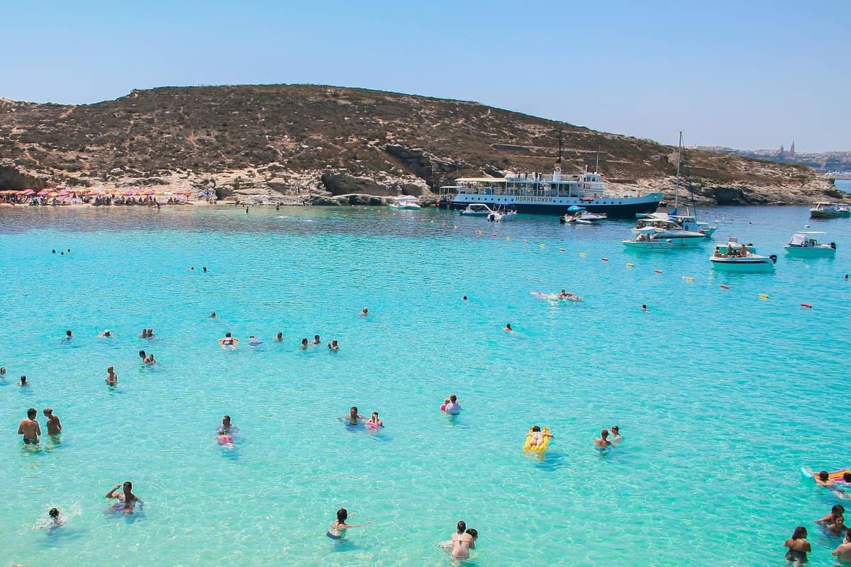 Folle di persone che nuotano nelle acque turchesi cristalline della Blue Lagoon, Comino, con barche e colline rocciose sullo sfondo.