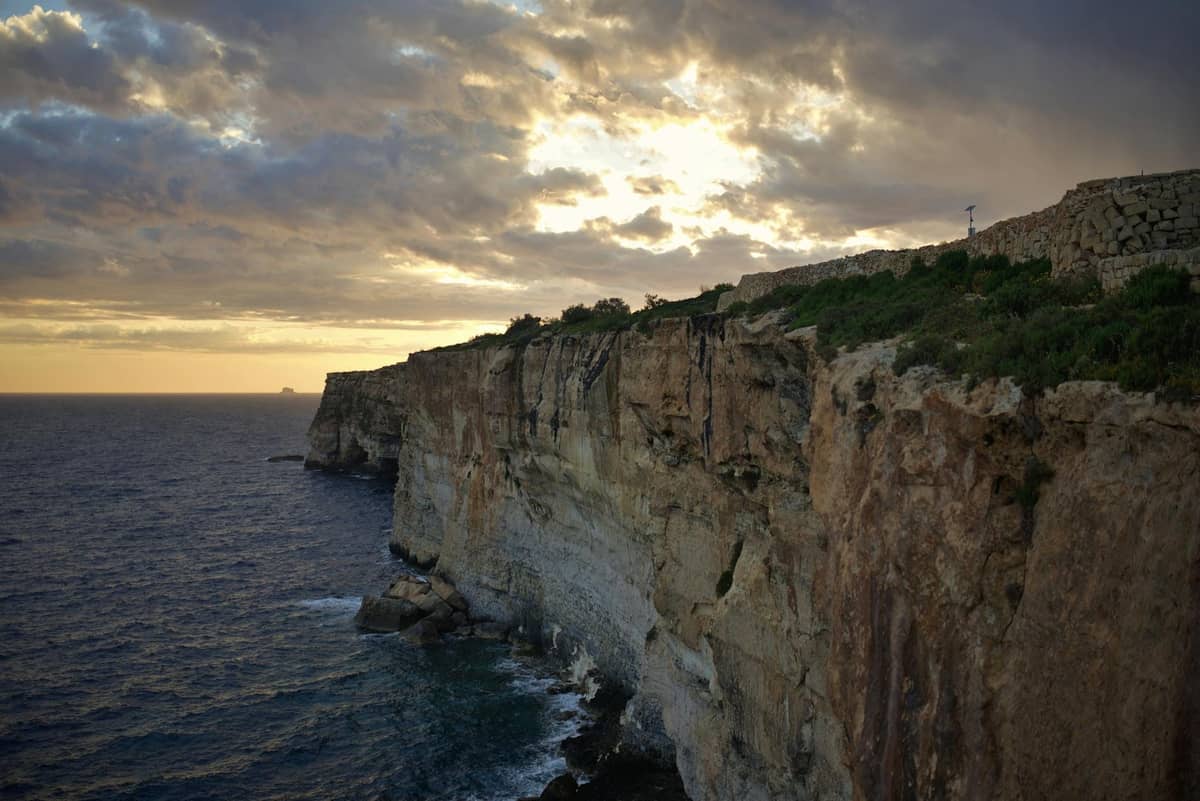 Drammatiche scogliere a picco sul mare nella luce dorata con le onde che si infrangono sotto sulla rocciosa costa maltese sotto un cielo colorato.