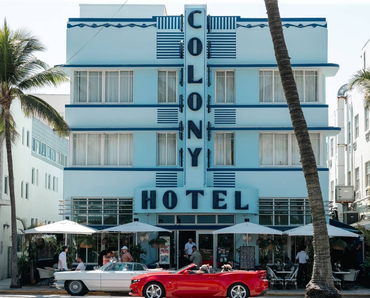 Art Deco Colony Hotel with classic cars and outdoor dining on Miami's famous Ocean Drive in bright daylight.