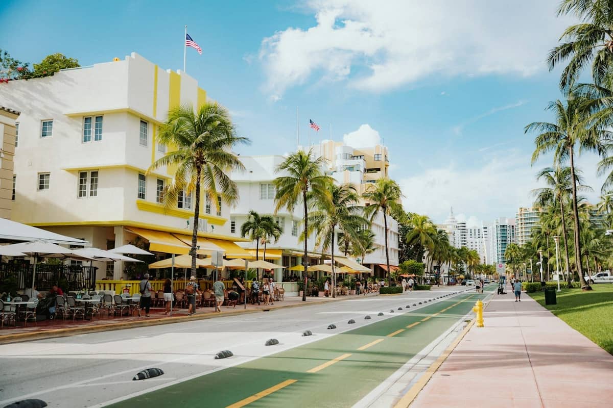 Bright and sunny view of Ocean Drive in South Beach, Miami, with art deco buildings, sidewalk cafes, palm trees, and people enjoying the outdoor atmosphere.