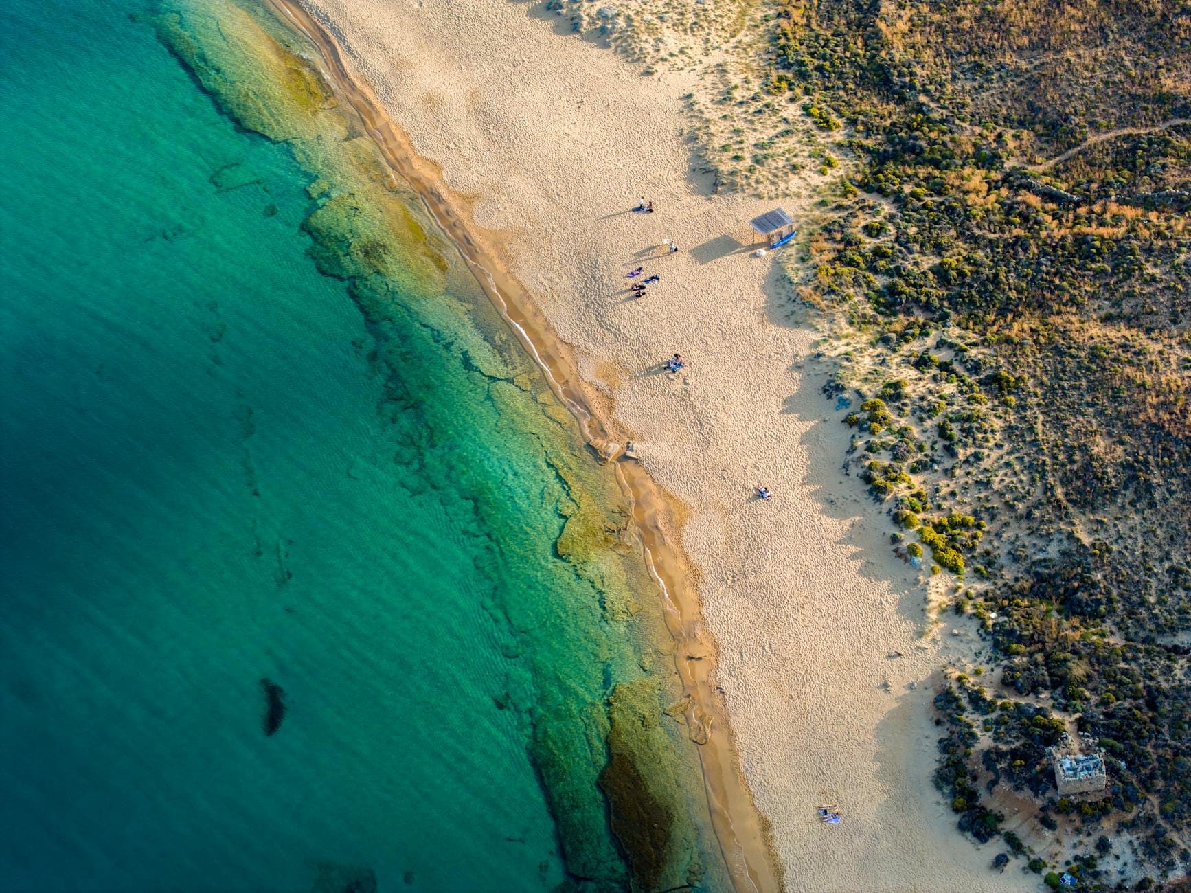 Vista aérea de una playa de arena tranquila y sin urbanizar con agua turquesa cristalina y escasos visitantes, rodeada de paisaje natural sin edificios.