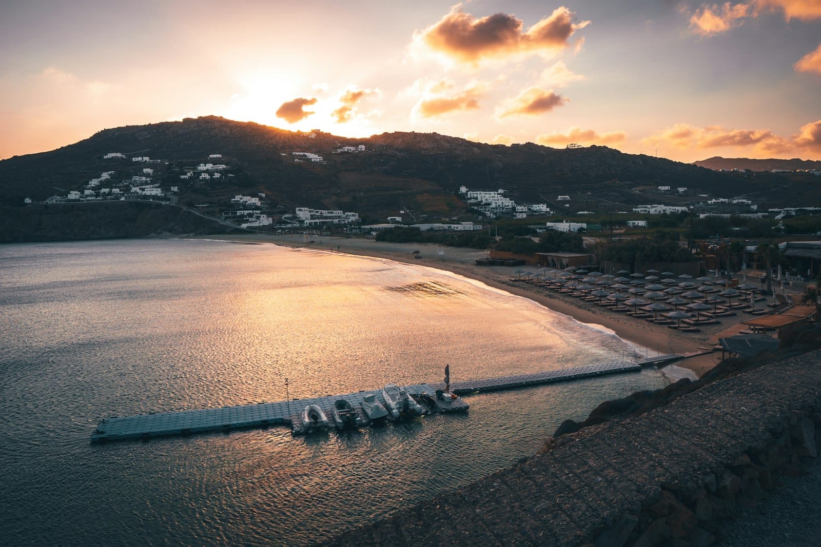 Amplia playa de arena en Mykonos con tumbonas, un largo muelle con botes y colinas al fondo al atardecer.