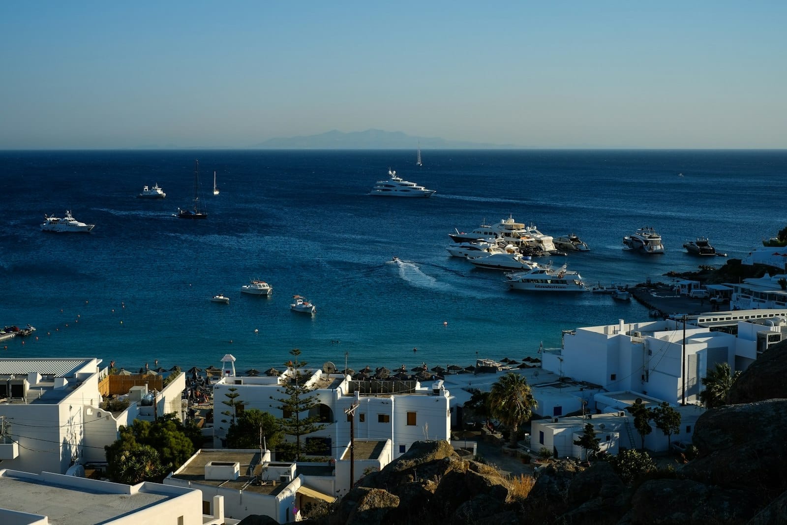 Yates y barcos anclados cerca de una playa de arena en Mykonos, con edificios blancos y mar azul bajo un cielo despejado.