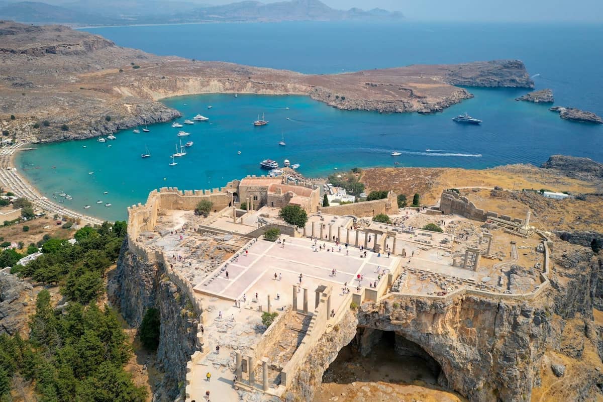 Aerial view of Lindos Acropolis perched on a cliff above turquoise bay, with dramatic coastline and moored boats in Rhodes, Greece.