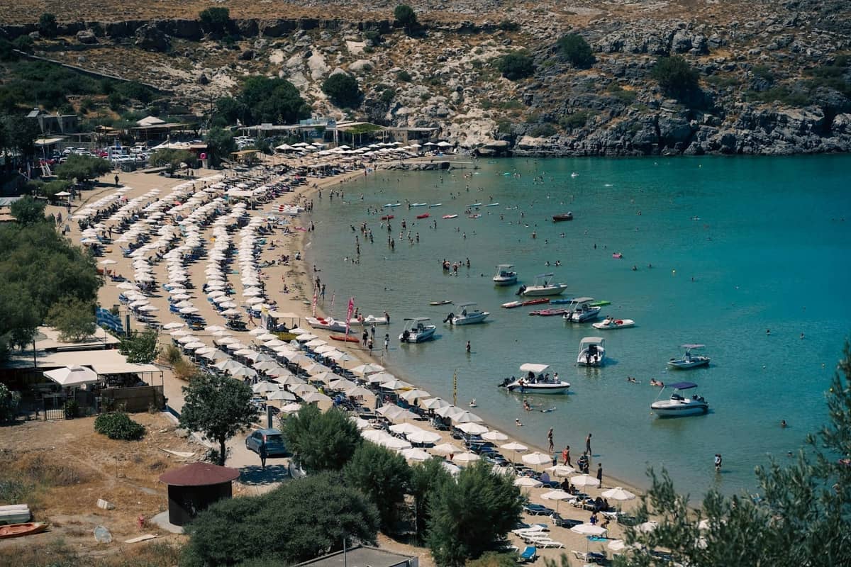 Aerial view of a sandy beach in Rhodes lined with umbrellas, shallow turquoise water, and families enjoying the sea with boats nearby.