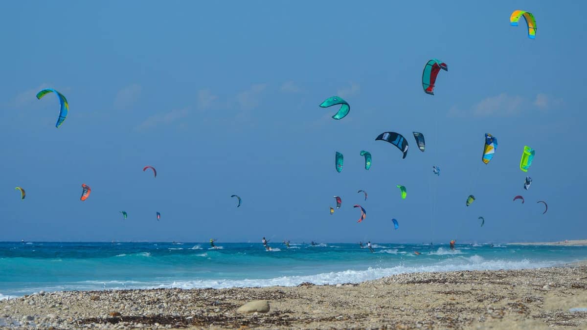 Pebbled beach in Rhodes with many colorful kitesurf sails in the sky above turquoise waves under a clear blue sky.