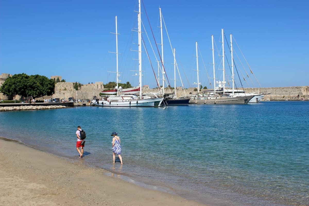Small sandy beach with two people walking, clear blue water, yachts, and medieval fortifications in the background in Rhodes.