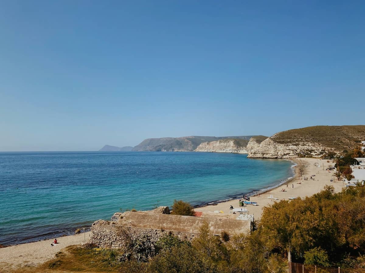 Wide sandy beach along a quiet coastline with clear blue water, sparse visitors, and rocky cliffs in the distance.