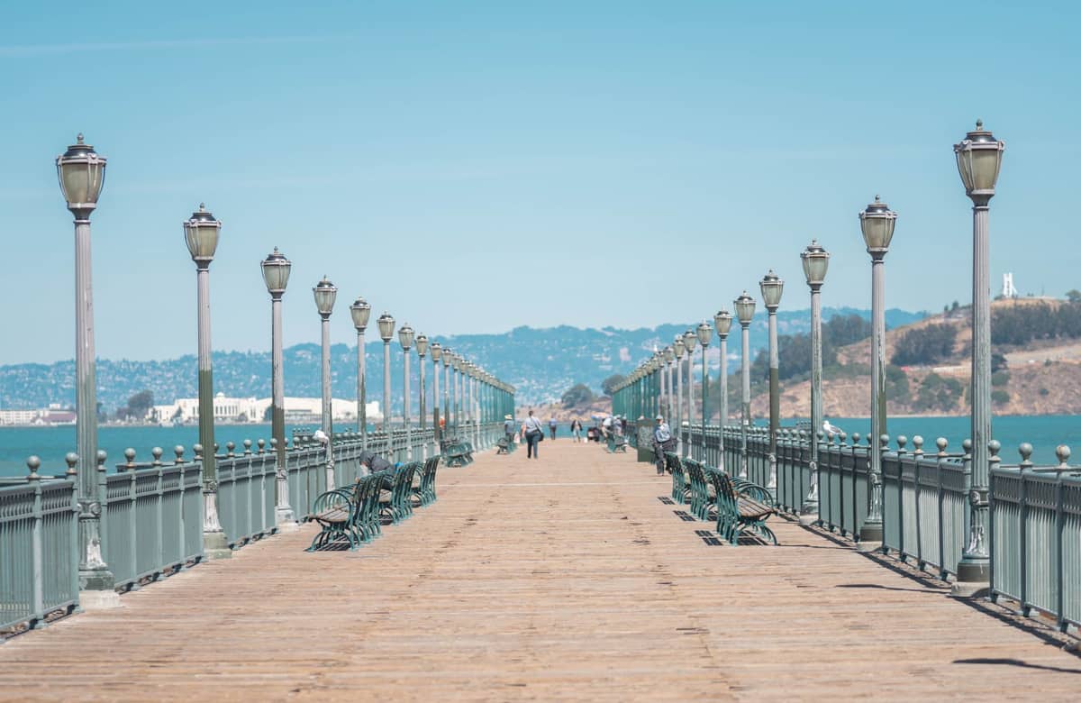 Lungo molo in legno con panchine e lampioni che si estende sulla Baia di San Francisco, persone che camminano e acqua visibile su entrambi i lati.