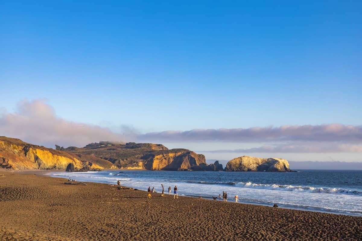 Vista panorámica de Baker Beach en San Francisco con personas en la arena, acantilados costeros y el océano Pacífico al fondo bajo un cielo azul despejado.