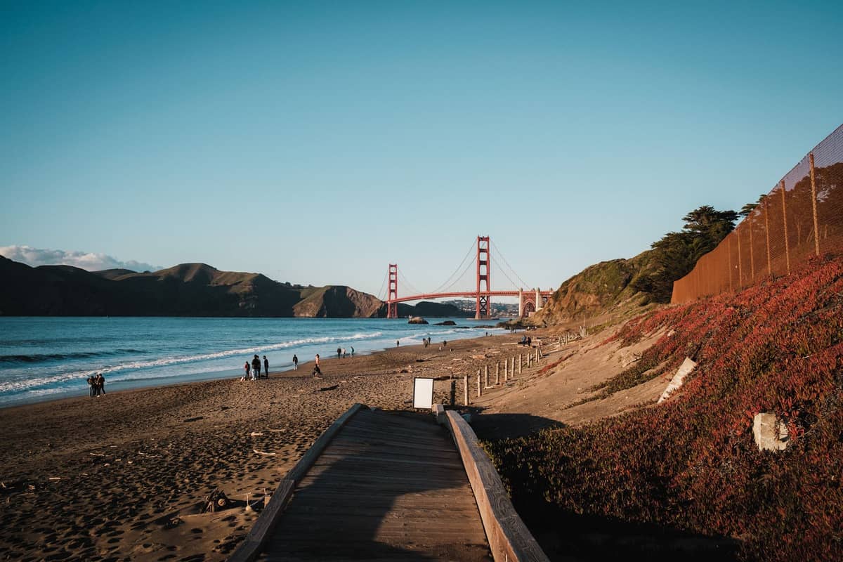 Ampia spiaggia sabbiosa con persone che passeggiano, una passeggiata sul lungomare e il Golden Gate Bridge sullo sfondo in una giornata serena.