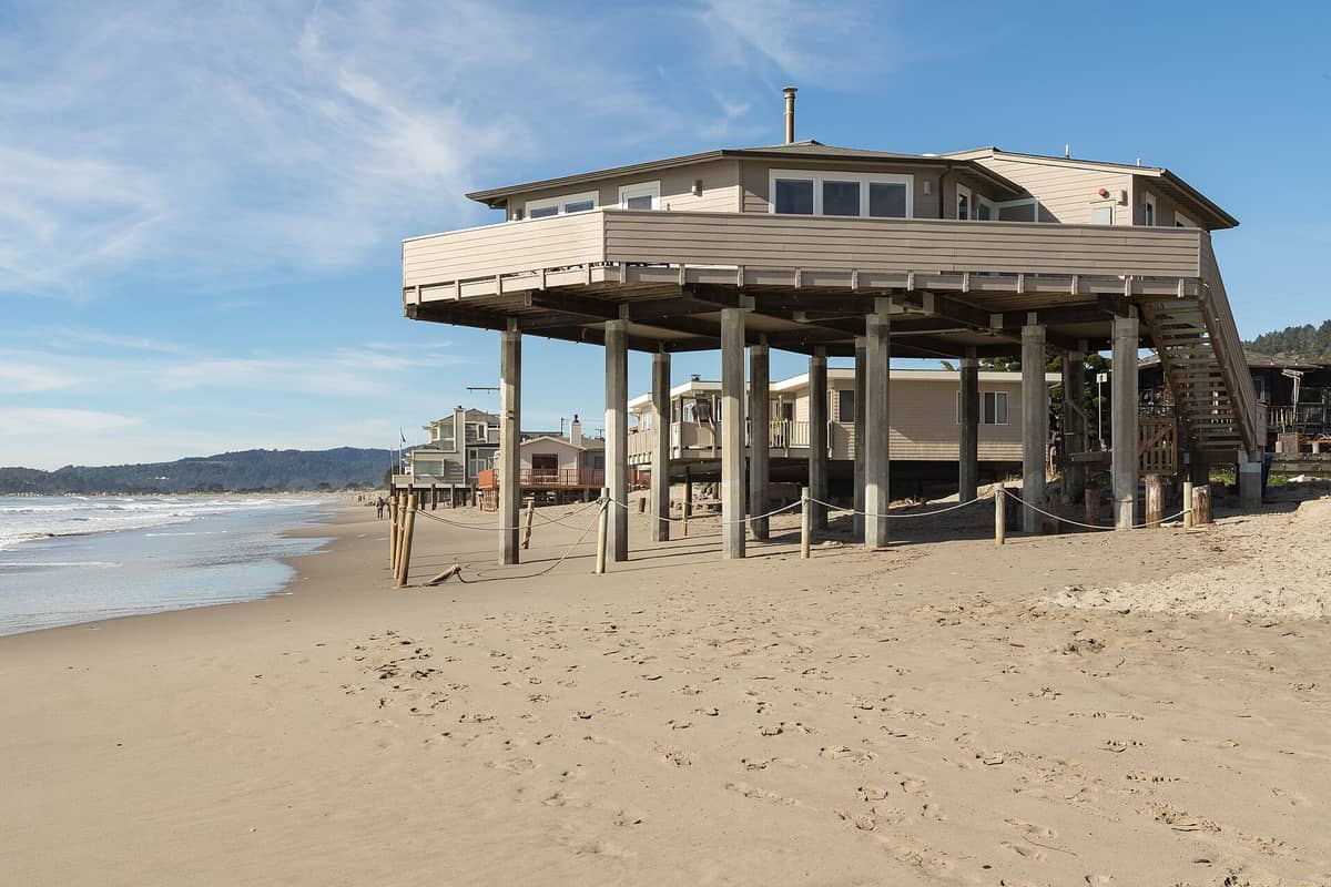 Ampia spiaggia sabbiosa con case su palafitte in primo piano e onde lungo la costa della California settentrionale sotto un cielo azzurro.