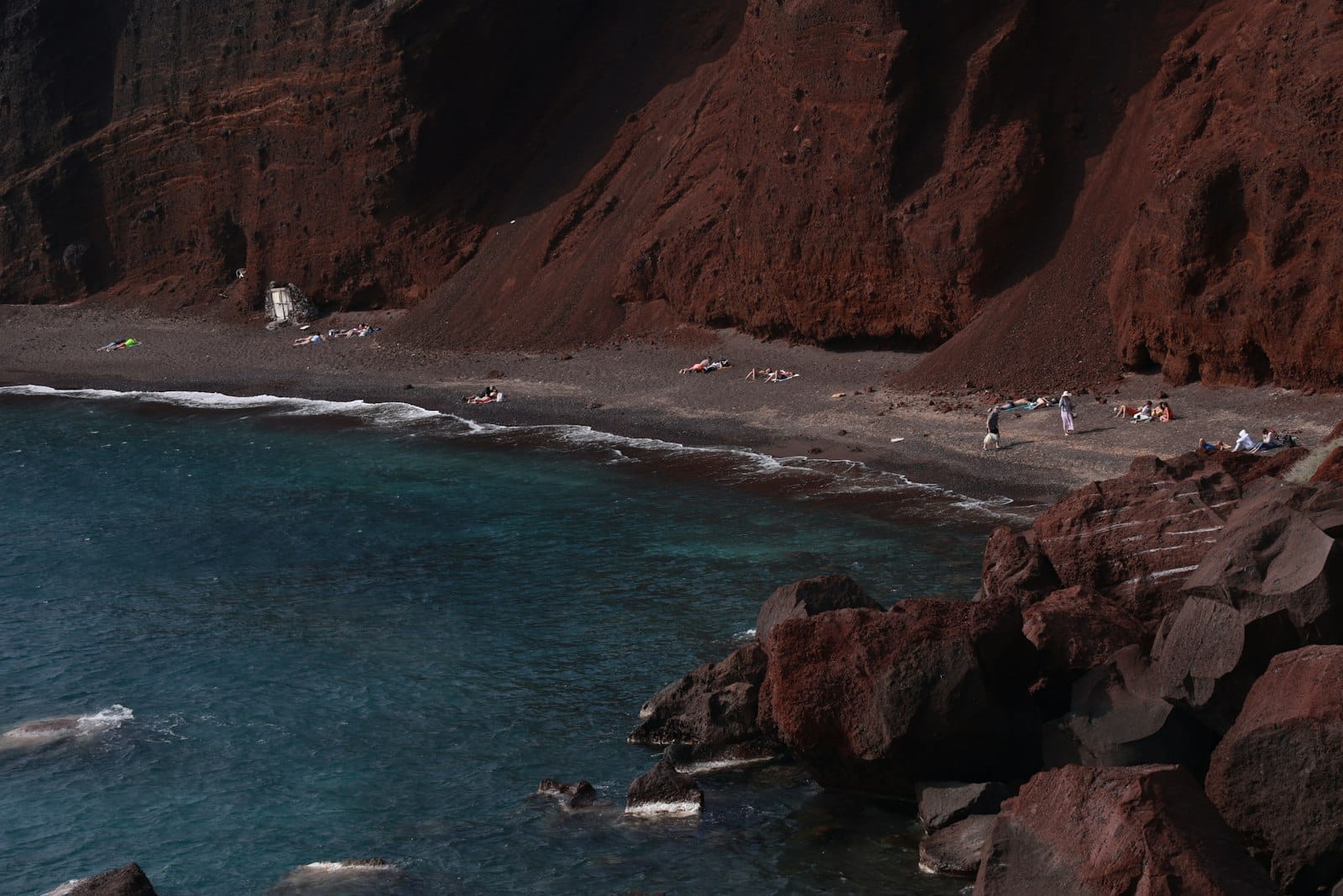 Vista impressionante da Praia Vermelha de Santorini com falésias vulcânicas vermelhas, areia negra e água azul profunda, emoldurada por rochas e banhistas ao longo da orla.