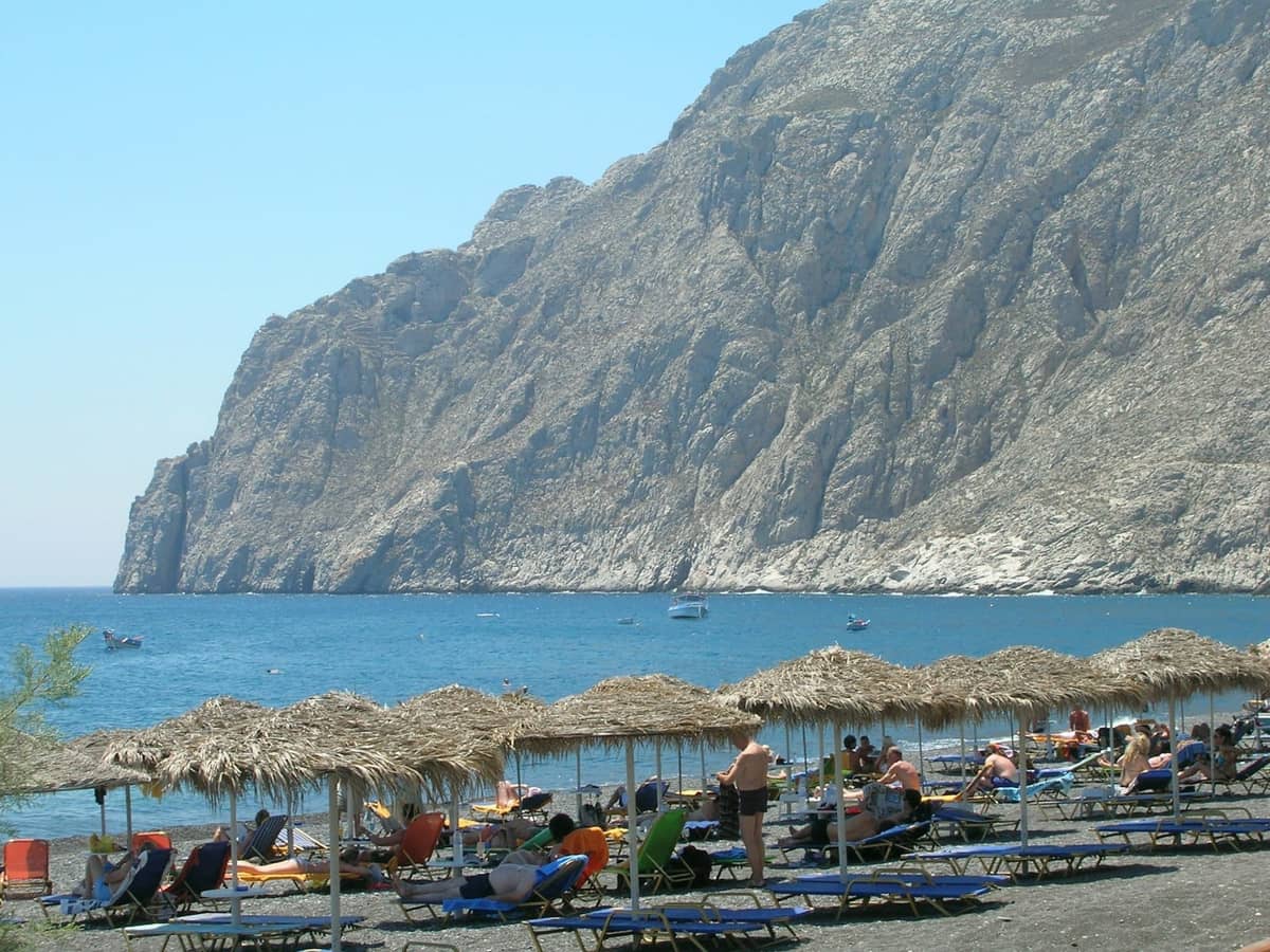 File di lettini e ombrelloni di paglia su una spiaggia di sabbia nera con un grande promontorio roccioso sullo sfondo.