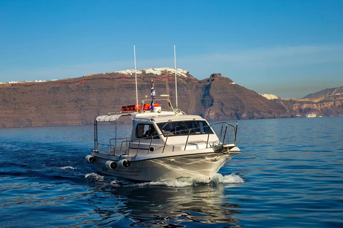 Piccola barca turistica che naviga su acque calme e blu con le scogliere della caldera di Santorini e gli edifici bianchi sullo sfondo sotto un cielo limpido.