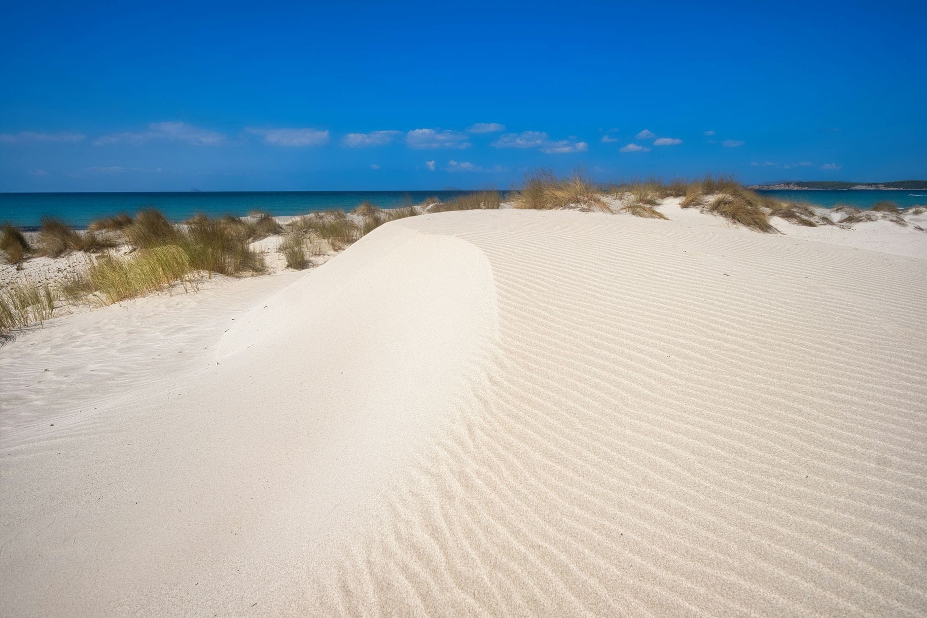 Weiße Sanddüne mit sanften Rücken und spärlichem Gras, türkisfarbenes Meer und strahlend blauer Himmel im Hintergrund.