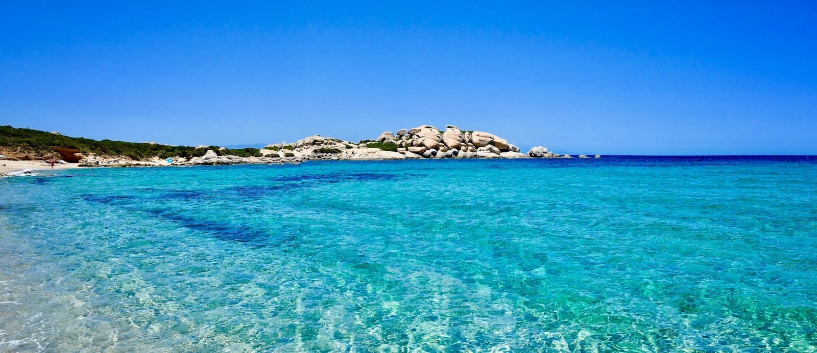 Weitläufige Landschaft eines sardischen Strandes mit kristallklarem türkisfarbenem Wasser, weißem Sand und felsigen Hügeln unter strahlend blauem Himmel – perfektes Titelbild für einen Reiseführer.