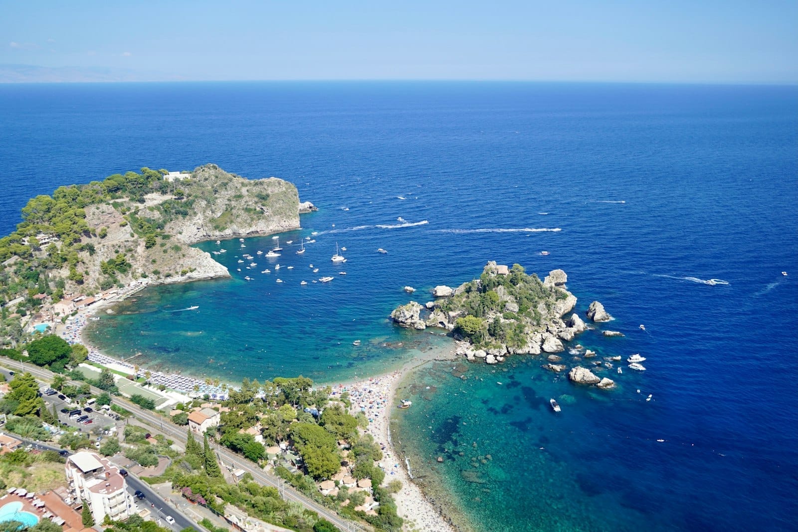 Luftaufnahme von Isola Bella und türkisfarbenem Wasser in der Nähe von Taormina, mit Stränden, Booten und grünen Hügeln entlang der ionischen Küste Siziliens.