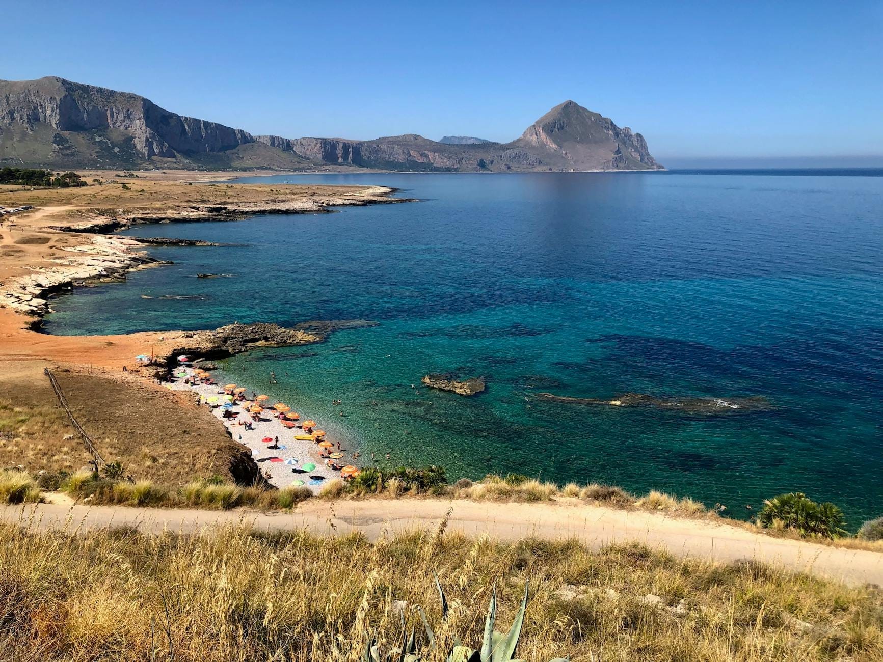 Dramatische Küstenlandschaft im Westen Siziliens mit felsigen Ufern, kristallklarem türkisfarbenem Wasser, einem Sandstrand mit Sonnenschirmen und Bergen in der Ferne.