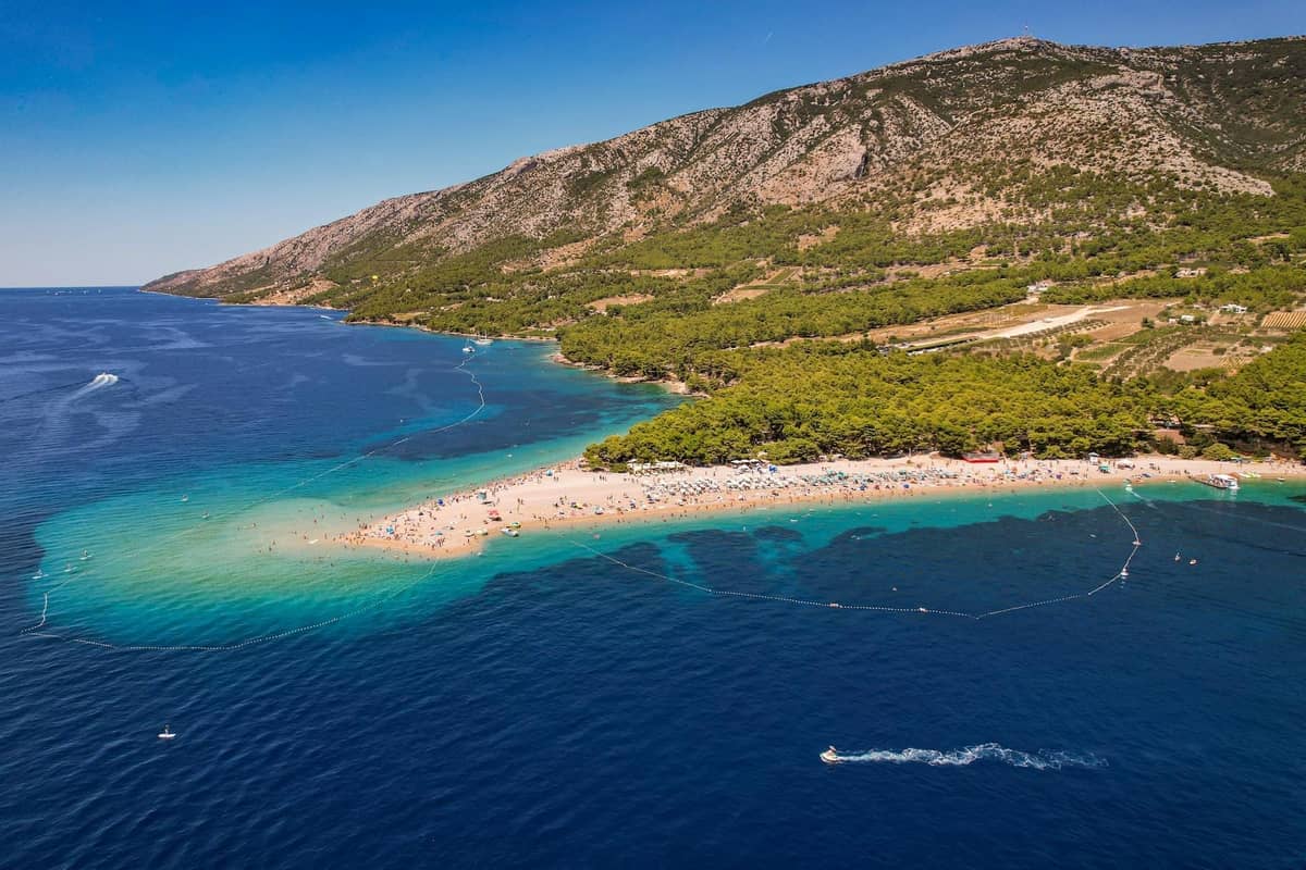 Luftaufnahme des Strandes Zlatni Rat auf der Insel Brač, mit türkisfarbenem Wasser, Pinienwald, Sandspitze und fernen Bergen unter blauem Himmel.