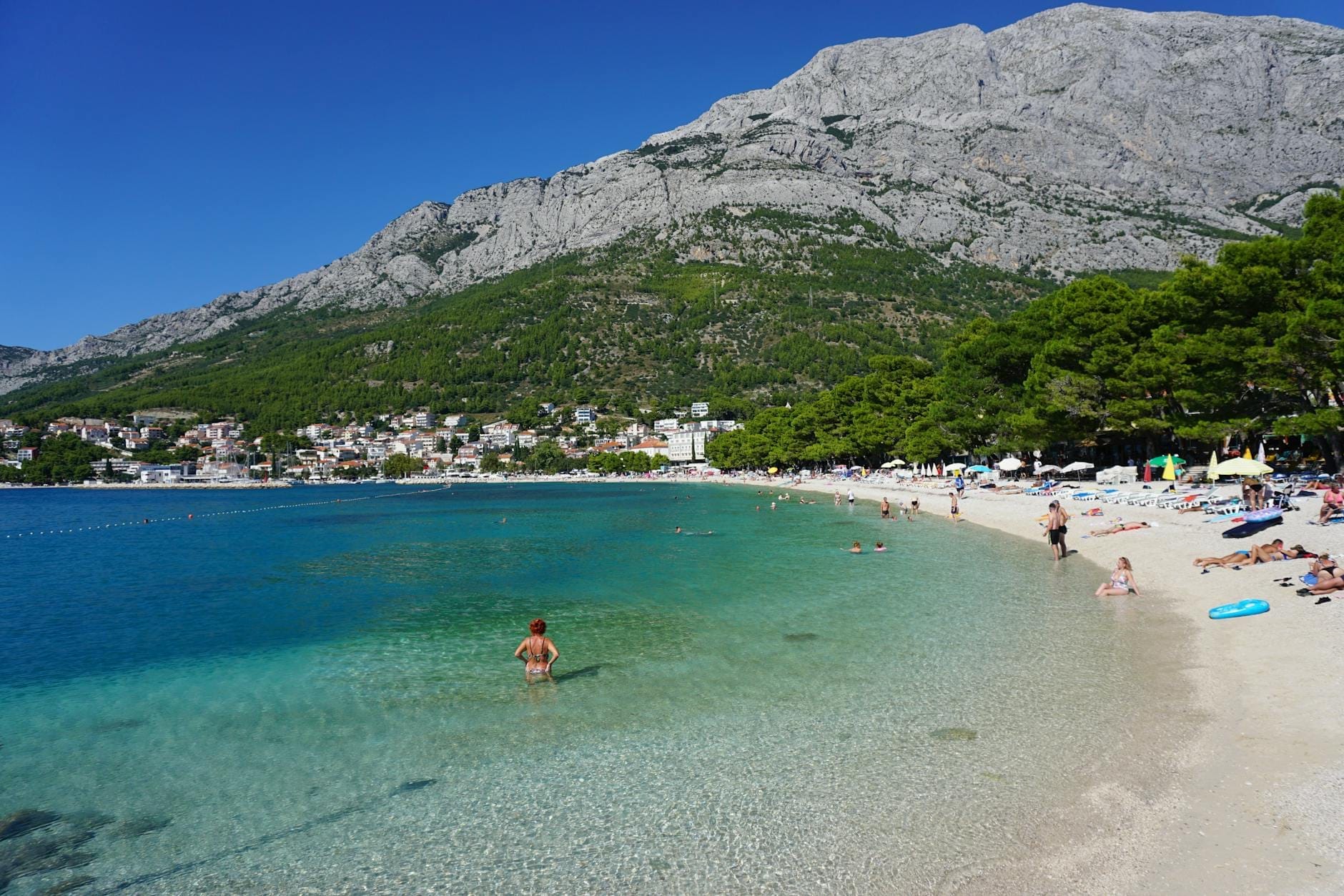 Banhistas e nadadores aproveitam uma praia de seixos cristalina perto de Split, com água turquesa, árvores verdes e montanhas rochosas ao fundo.