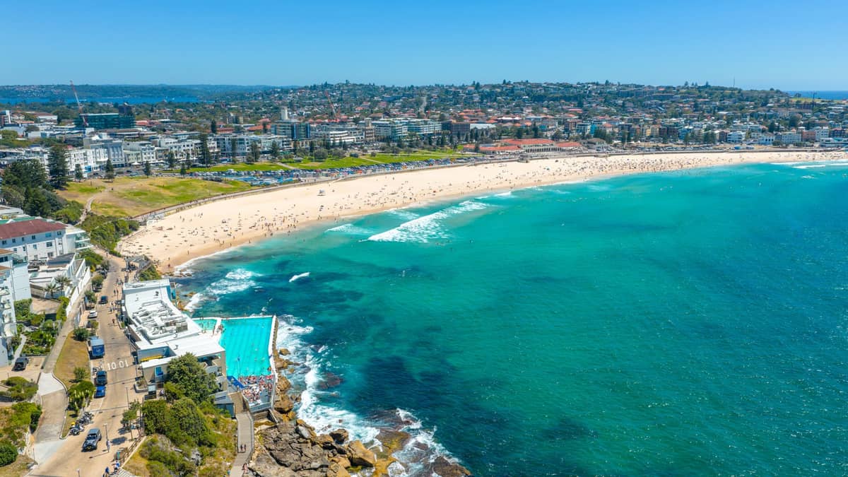 Vista aérea de Bondi Beach, con oleaje turquesa, orilla de arena, piscina de Bondi Icebergs y los suburbios del este de Sydney bajo un cielo azul despejado
