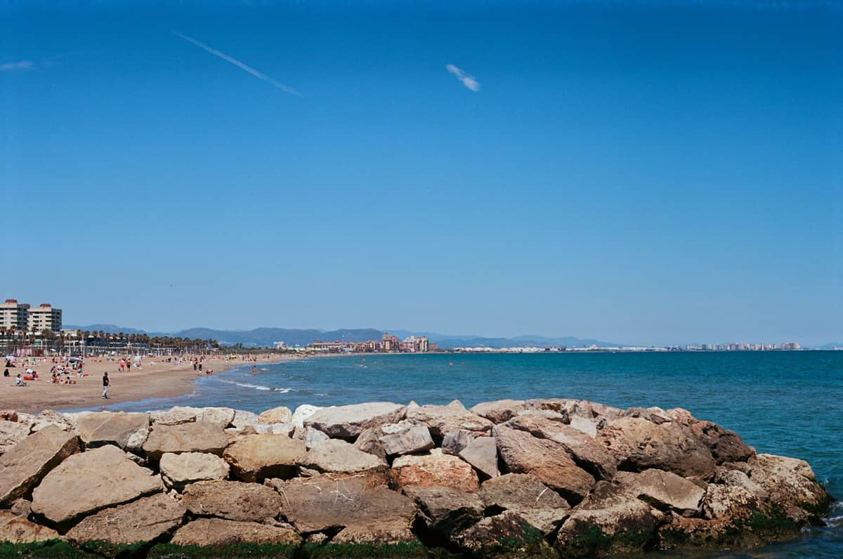 Vista sulla spiaggia urbana Malvarrosa di Valencia con edifici sul lungomare, sabbia dorata, molo roccioso e Mar Mediterraneo sotto un cielo azzurro.