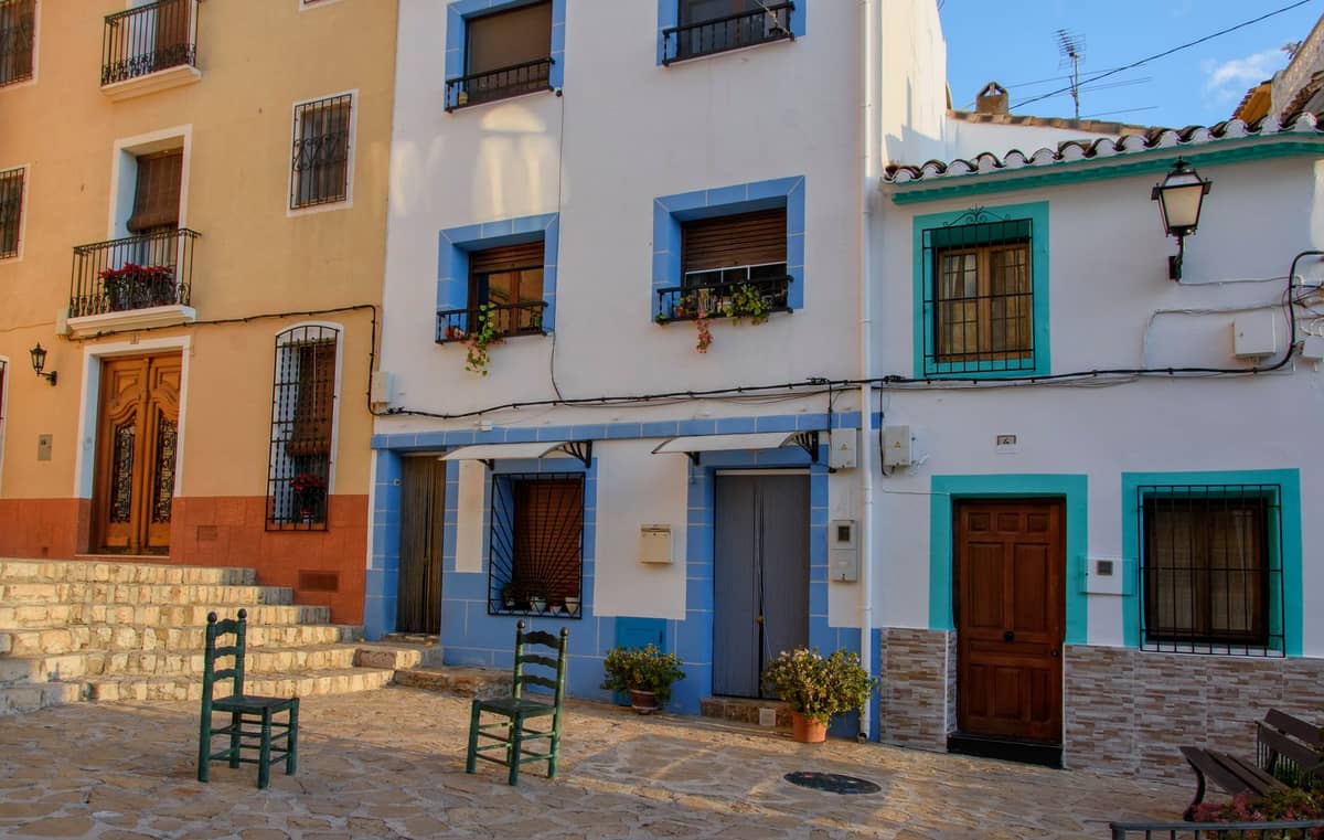 Traditionelle bunte Häuser mit blauen Rahmen und zwei leeren Stühlen auf einem sonnigen Platz in einem spanischen Küstenviertel.