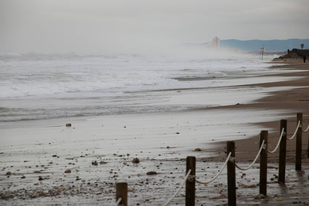 Wilde Sandstrand mit vereinzelten Kieselsteinen und Holzpfosten mit Seil, heranrollende Wellen und dunstiger Horizont in der Ferne.