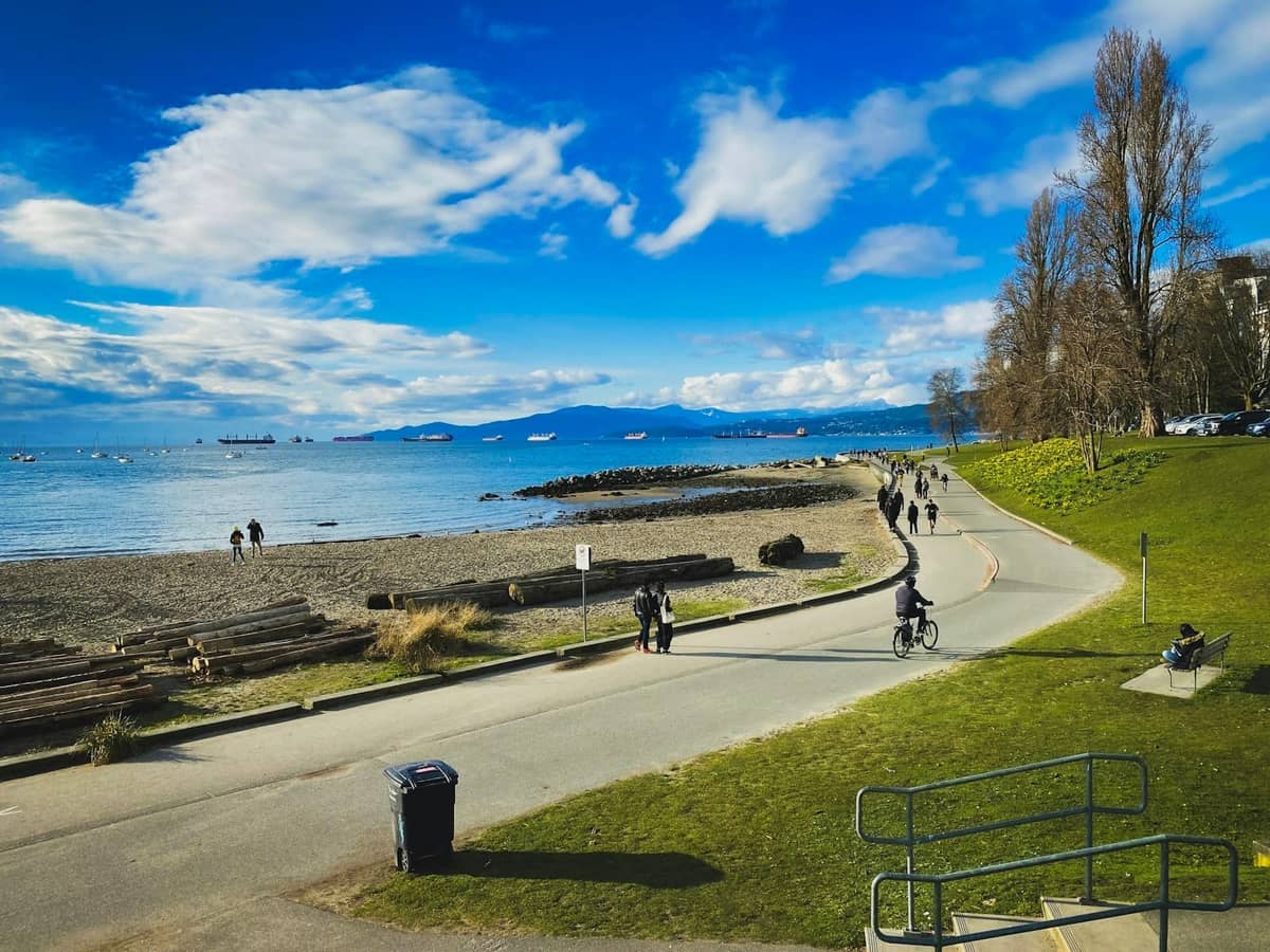 Vancouver Seawall ที่คึกคักด้วยผู้คนปั่นจักรยานและเดินเล่นริมชายหาด ท่อนซุง มหาสมุทร และวิวภูเขาอันกว้างไกลใต้ท้องฟ้าสดใส