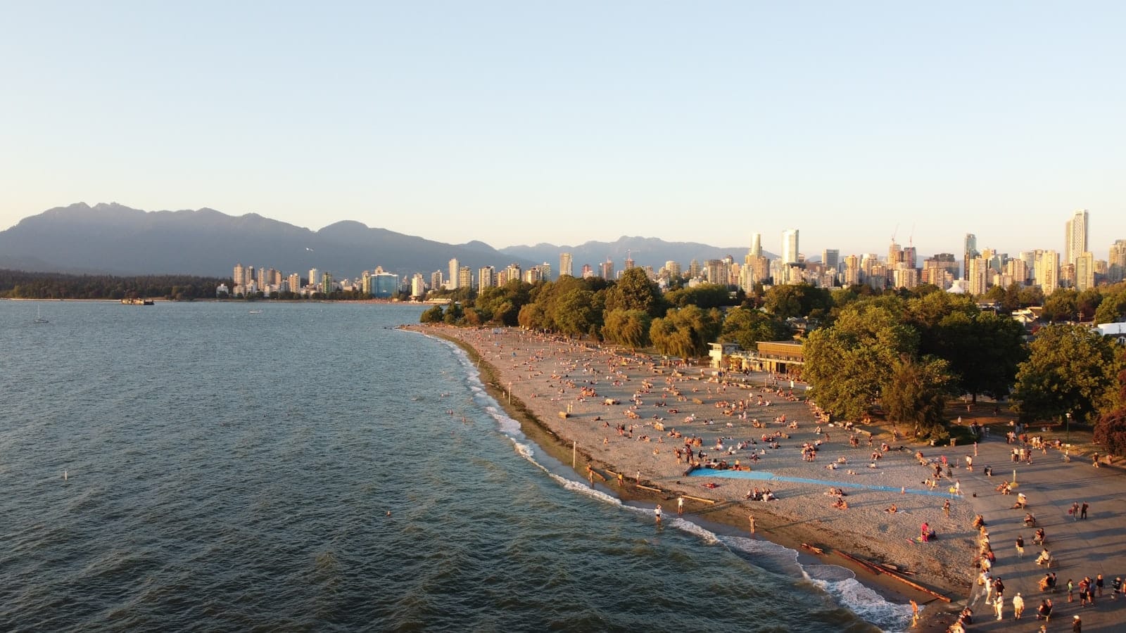 Uma ampla praia de areia em Vancouver com pessoas relaxando, montanhas ao fundo e o skyline da cidade sob um céu limpo ao entardecer.